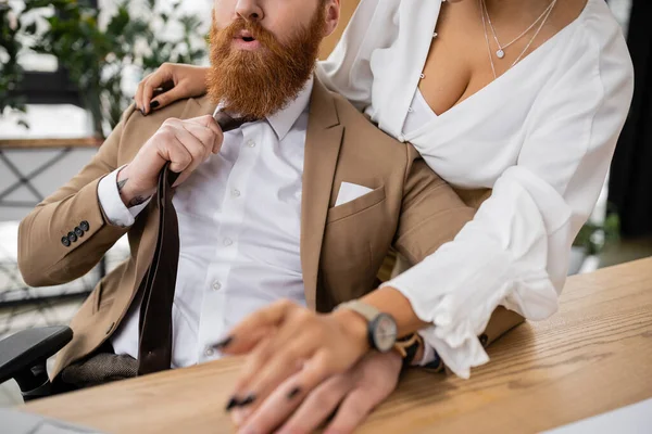 Cropped view of seductive african american woman leaning on bearded businessman pulling tie in office — Stock Photo