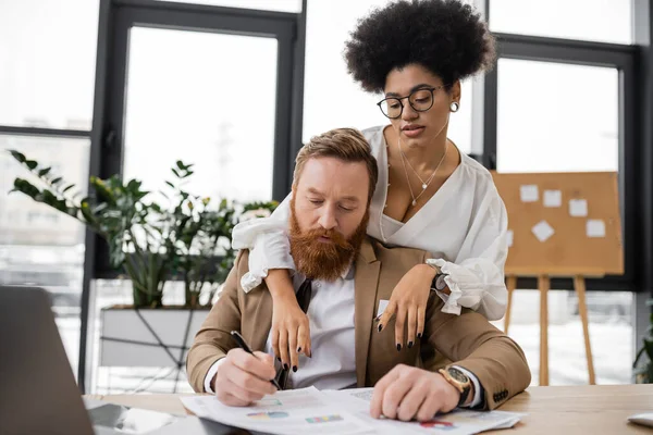 Seductive african american woman leaning on shoulders of bearded man while pointing at charts — Photo de stock