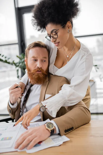 Passionate african american woman leaning on shoulders of bearded man while pointing at charts — Stock Photo