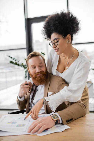 Passionate african american woman leaning on shoulders of cheerful man while pointing at charts — Stock Photo
