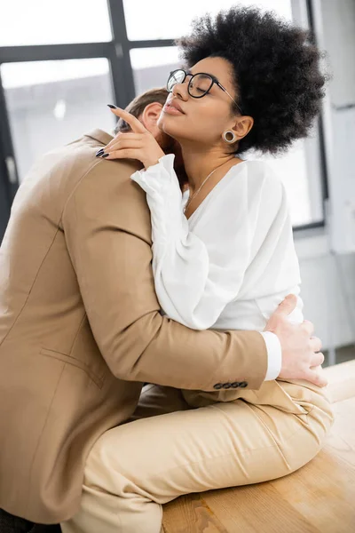 Passionate man kissing neck of african american woman in glasses sitting on desk in office — Photo de stock