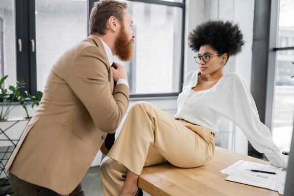 African american woman sitting on office desk and seducing bearded man pulling tie — Foto stock