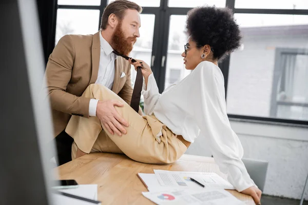 Side view of african american woman pulling tie of bearded man sitting on office desk — Stock Photo