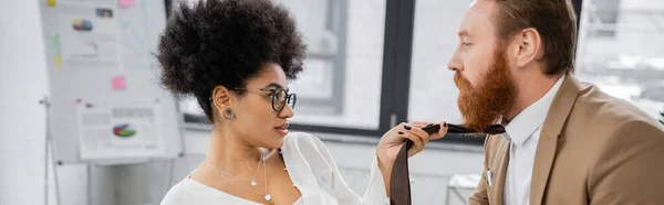 Side view of african american woman pulling tie of bearded coworker in office, banner — Stock Photo
