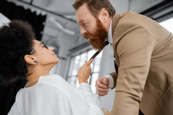 Side view of smiling african american woman pulling tie of bearded coworker in office - foto de stock