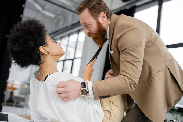 Side view of curly african american woman pulling tie of bearded coworker while lying on office table — Stock Photo