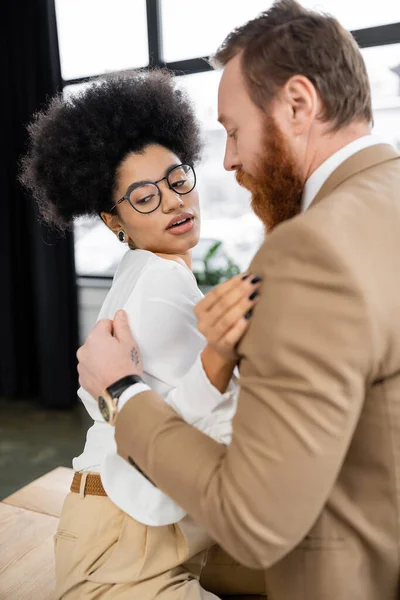 Tattooed man touching shirt while undressing curly african american woman in office — Stock Photo