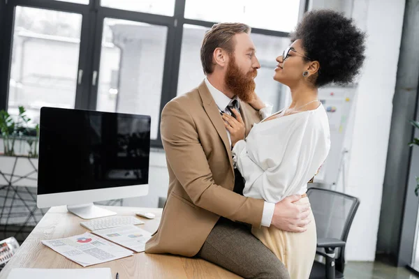 Brunette african american woman seducing bearded businessman in suit sitting on desk — Stock Photo