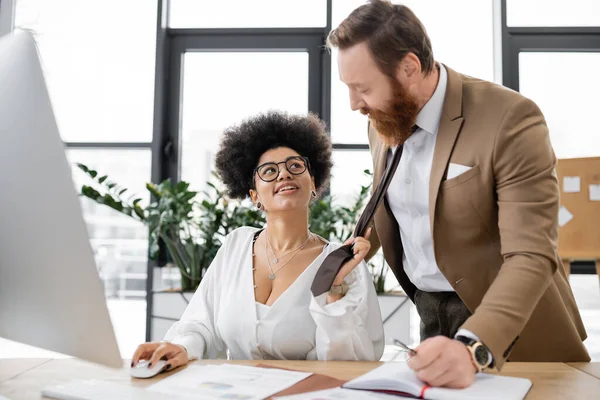 Smiling african american woman pulling tie of bearded businessman in office — Stock Photo