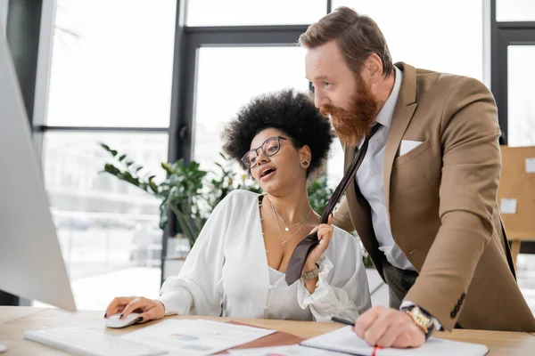 Cheerful african american woman pulling tie of bearded businessman in office — Stock Photo
