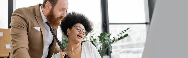 Young african american woman pulling tie of bearded businessman in office, banner — Foto stock