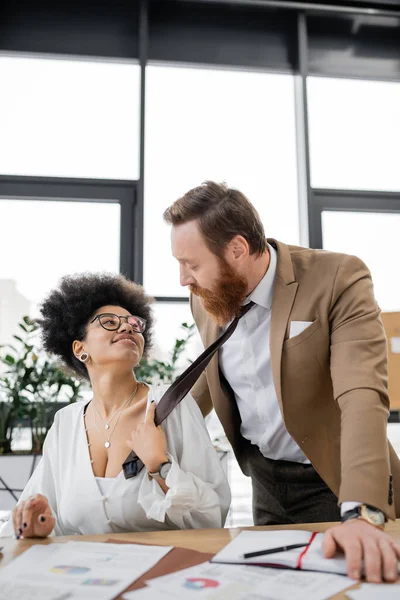 Smiling african american woman pulling tie of bearded businessman in office — Foto stock