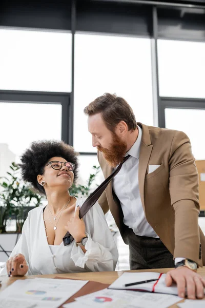 Smiling african american woman pulling tie of bearded businessman in office - foto de stock