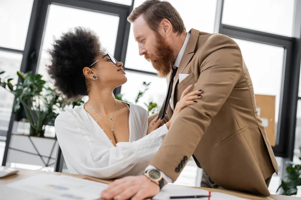 Smiling african american woman pulling tie of passionate businessman in office — Fotografia de Stock