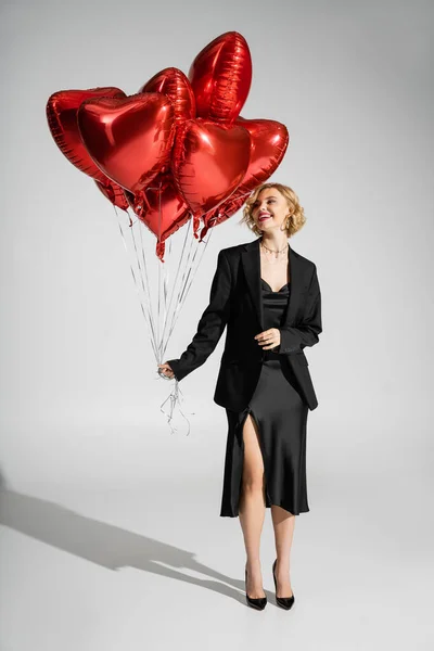 Full length of cheerful woman in black slip dress and blazer looking at red heart-shaped balloons on grey — Stock Photo