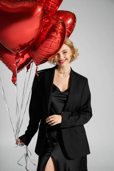 Счастливая молодая женщина в черном платье и блейзере с красными шарами в форме сердца, изолированными на сером — стоковое фото