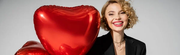 Счастливая молодая женщина в черном блейзере улыбается рядом с красными шарами в форме сердца, изолированными на сером баннере — стоковое фото