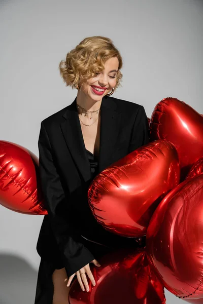 Positive blonde woman in black blazer and slip dress standing with red balloons isolated on grey — Stockfoto