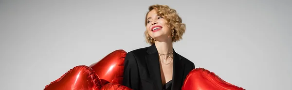 Positive woman in black blazer standing with red heart-shaped balloons isolated on grey, banner — Stock Photo