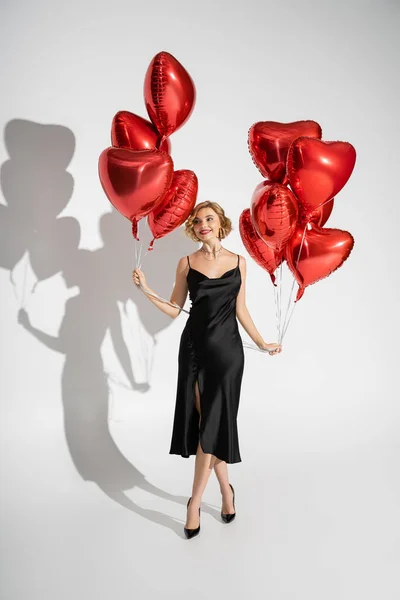 Full length of smiling woman in satin slip dress standing with red heart-shaped balloons on grey — Stockfoto