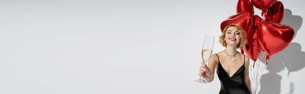 Happy woman in black slip dress holding glass of champagne and standing with red balloons on grey, banner — Stock Photo