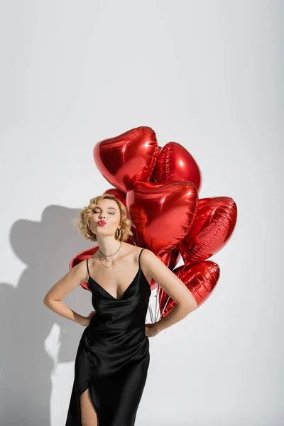 Blonde woman in black slip dress pouting lips near red heart-shaped balloons on grey — Photo de stock