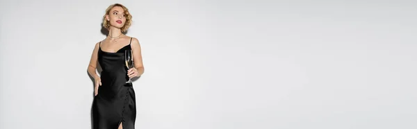 Blonde woman in satin slip dress holding glass of champagne while standing with hand on hip isolated on grey, banner — Stock Photo