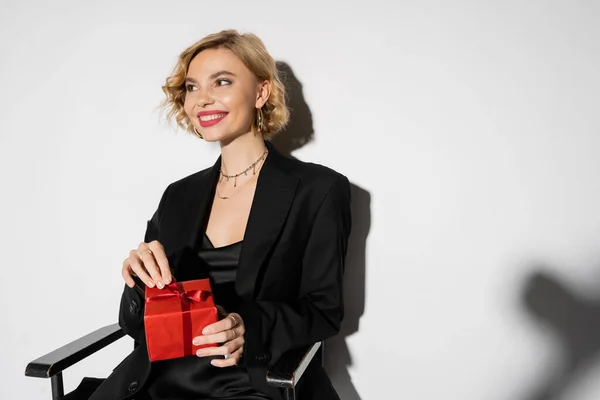 Positive woman in black blazer and slip dress holding wrapped present while sitting on grey — Stock Photo