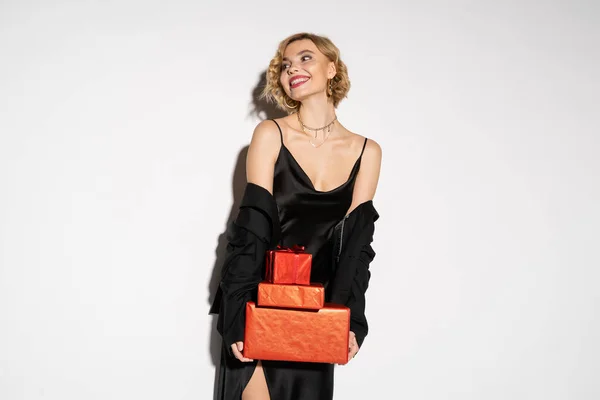 Pleased woman in black blazer and satin slip dress holding wrapped red presents on grey background — Stock Photo