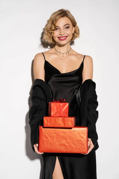 Positive woman in black blazer and satin slip dress holding wrapped red presents on grey — Stockfoto