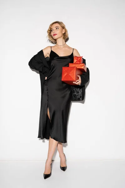Blonde woman in black blazer and satin slip dress holding wrapped red presents while posing with hand on hip on grey — Photo de stock