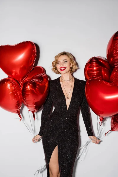 Happy and blonde woman in tight black dress holding red heart-shaped balloons on grey — Stockfoto
