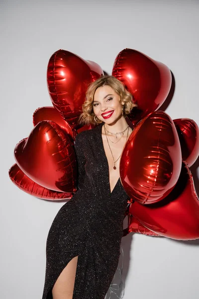 Joyful young woman in tight black dress holding red heart-shaped balloons on grey — Stock Photo