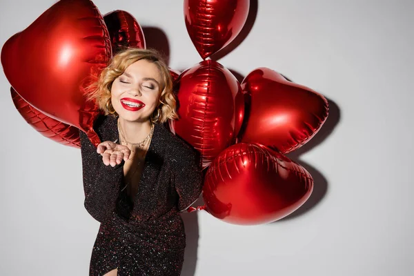 Happy and blonde woman in black dress holding red heart-shaped balloons and gesturing on grey — Stock Photo