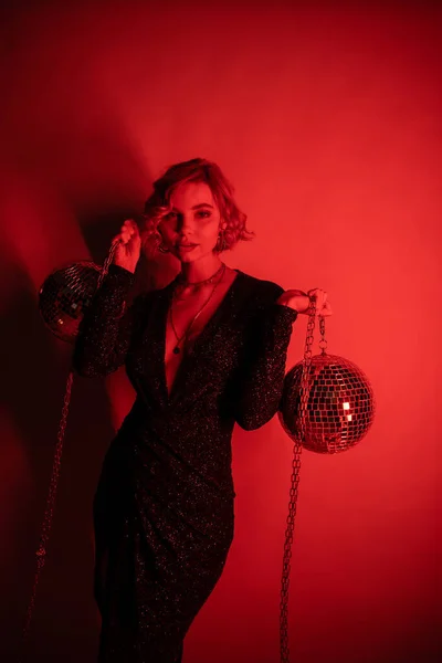 Young woman with blonde hair standing in black tight dress and holding chains with disco balls on red — Fotografia de Stock