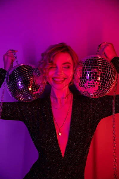 Cheerful young woman with blonde hair standing in black dress and holding chains with disco balls on purple and pink - foto de stock