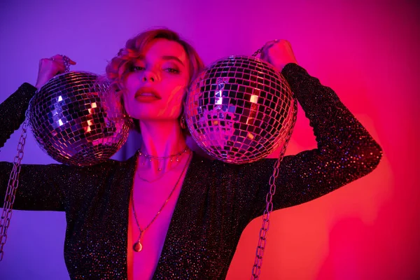 Young woman with blonde hair standing in black dress and holding chains with disco balls on purple and pink — Foto stock