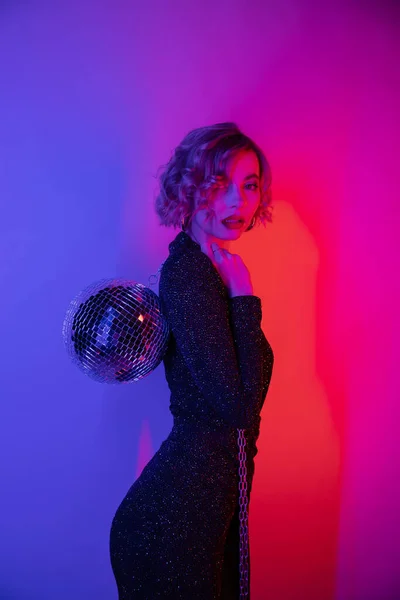 Young woman with blonde hair standing in tight dress and holding chain with disco ball on purple and pink — Fotografia de Stock