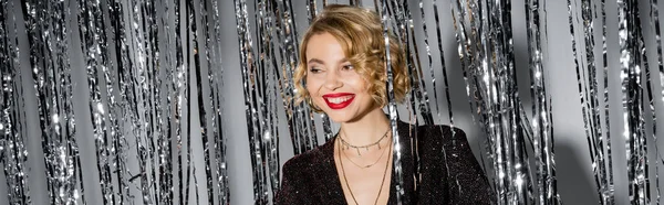 Positive young woman with red lips standing in black dress near shiny tinsel curtain on grey, banner — Foto stock