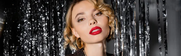 Blonde young woman with red lips looking at camera near shiny tinsel curtain on grey, banner — Fotografia de Stock