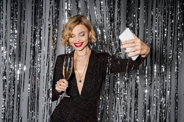 Cheerful woman in black tight dress taking selfie while holding glass of champagne near tinsel curtain on grey — Foto stock