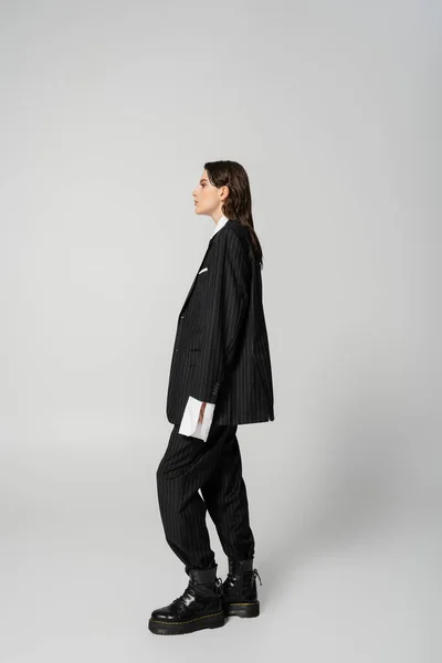Side view of trendy woman posing in black oversize pantsuit and rough boots on grey background — Foto stock