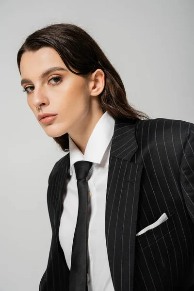 Young brunette woman in white shirt and black blazer with tie looking at camera isolated on grey — Stock Photo