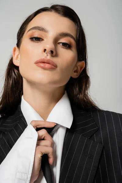 Portrait of trendy woman with makeup and piercing touching black tie and looking at camera isolated on grey — Stockfoto