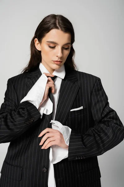 Fashionable young woman in oversize shirt and blazer adjusting tie isolated on grey — Stockfoto