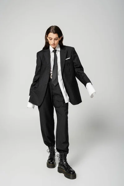 Полная длина стильная женщина в черном крупногабаритном костюме и грубых сапогах стоя на сером фоне — стоковое фото