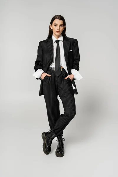 Full length of brunette woman in elegant oversize pantsuit and rough boots standing with hands in pockets on grey background — Stockfoto