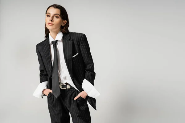 Pretty woman in black suit and white shirt standing with hands in pockets while looking at camera isolated on grey — Stock Photo