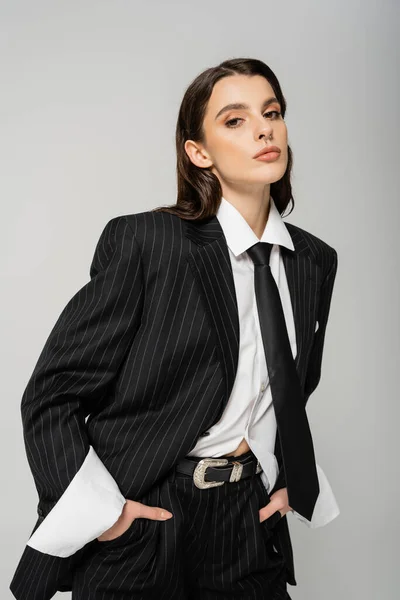 Pretty young woman in black stylish suit and white shirt looking at camera while posing with hands in pockets isolated on grey - foto de stock
