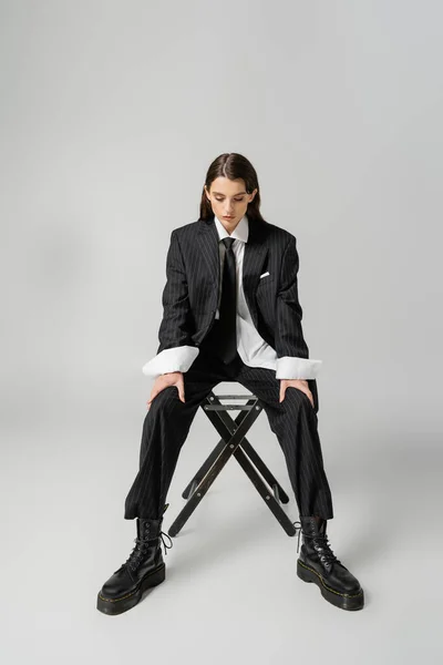 Полная длина красивой модели в черном элегантном костюме и грубых кожаных сапогах, сидящих на стуле на сером фоне — стоковое фото